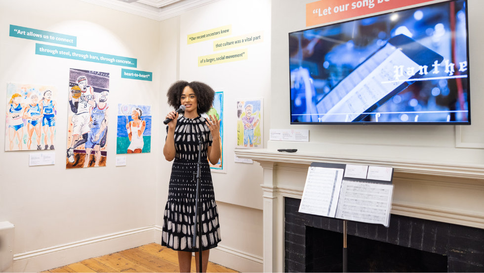 Curator Melaine Ferdinand-King '25 Ph.D speaks into a microphone at the exhibition opening.