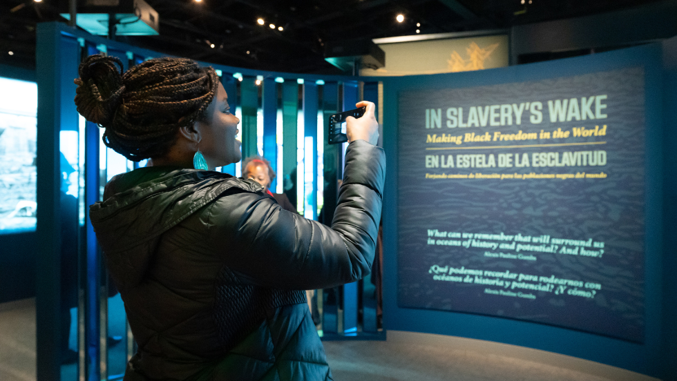 A woman takes an image of the exhibition's title wall reading "In Slavery's Wake: Making Black Freedom in the World".
