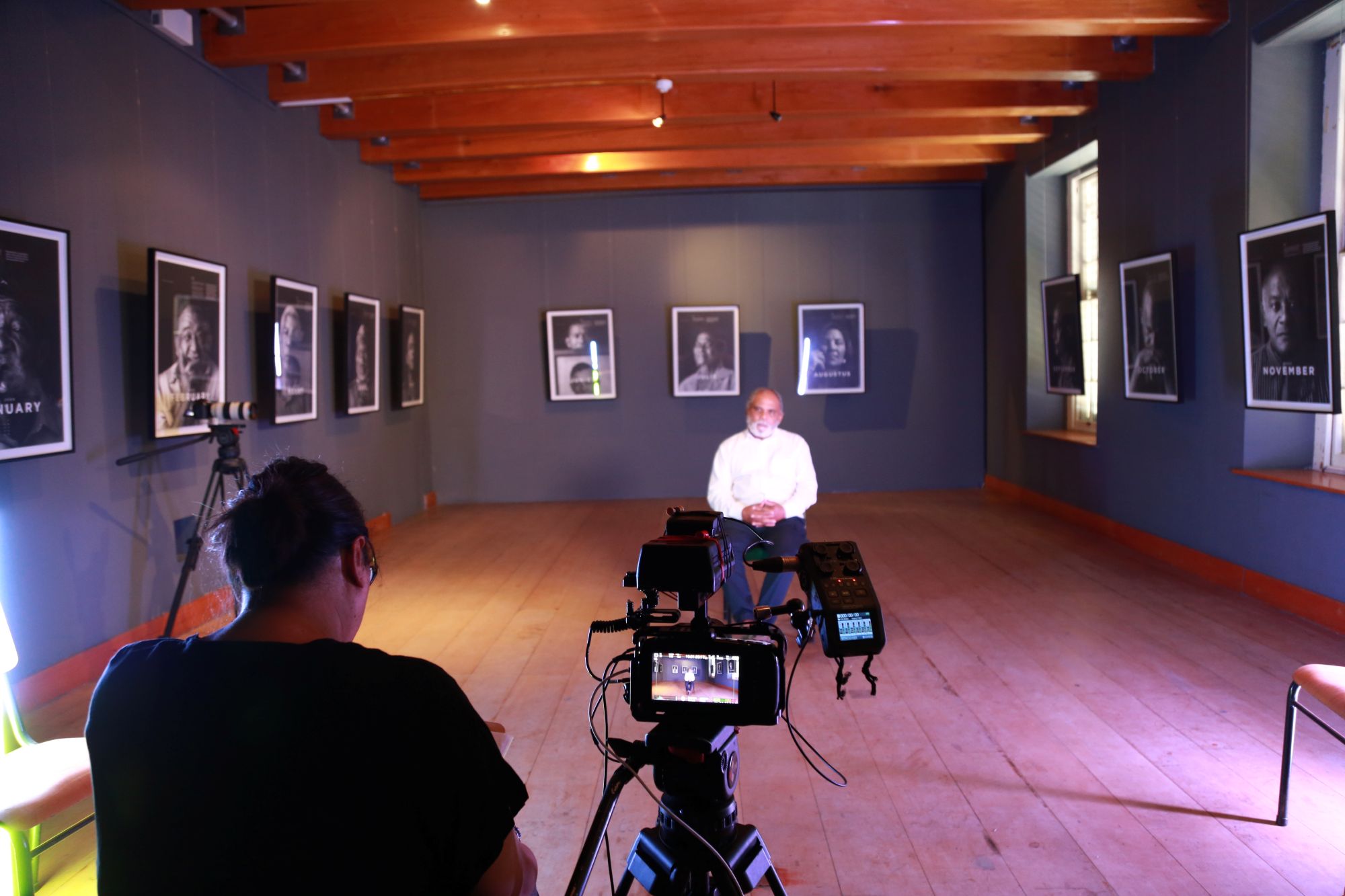 a man sits in the middle of a gallery of portraits while being interviewed and filmed