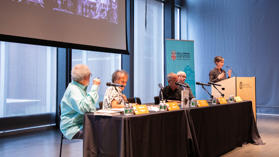 Prof. Brian Meeks, Prof. Lindsay Caplan, Prof. Geri Augusto, and Prof. Bogues sit in front of a projector screen while Prof. Philip Gould addresses the crowd from a podium next to them.