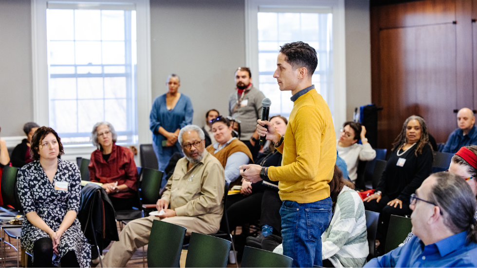 A man in a yellow sweater participates in a Q&A amidst an audience who gazes towards him.