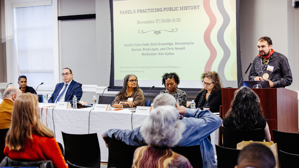 Panelist Brad Lopes speaks at the podium, fellow panelists sit a table next to the podium.