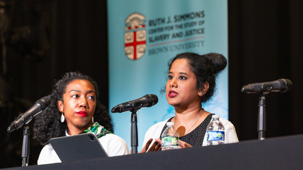 A woman sitting in front of a microphone stand addresses the audience while her fellow panelist glances over and listens in.