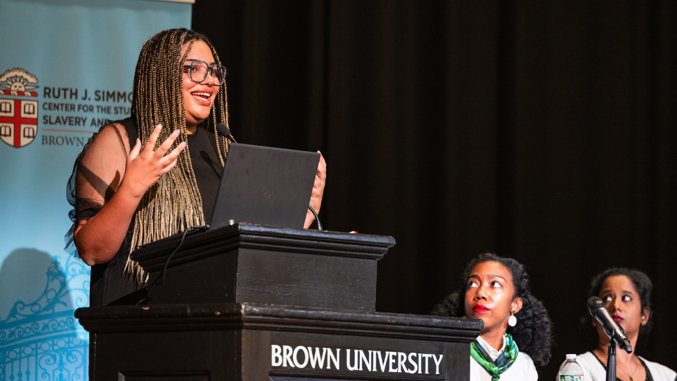 A speaker addresses the crowd at the podium, fellow panelists listen in.