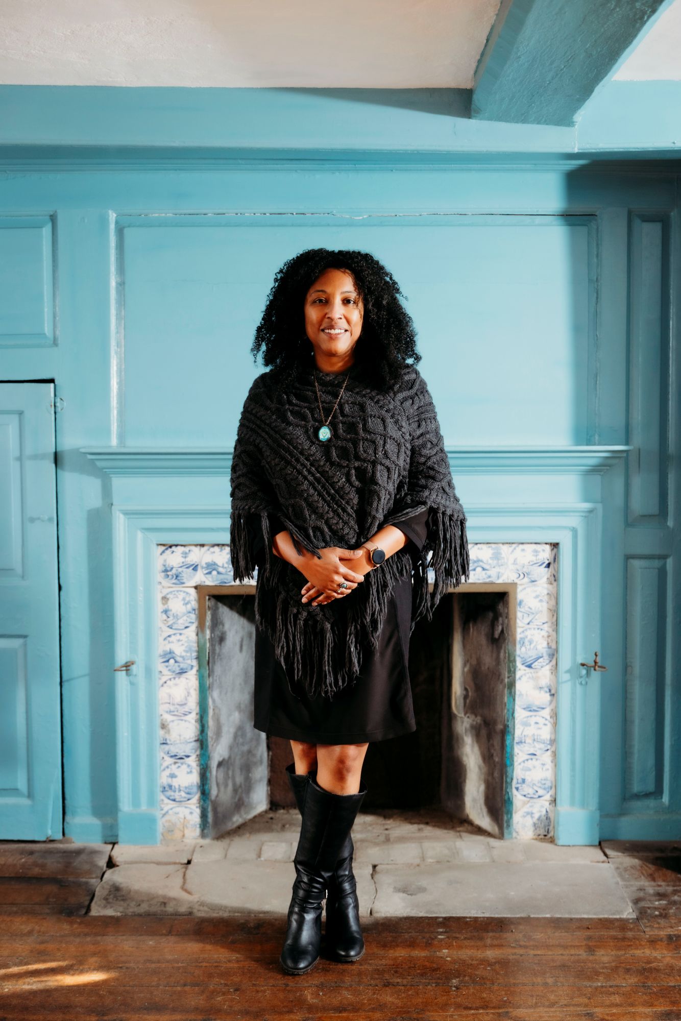 A woman stands in front of a fireplace in a colonial house
