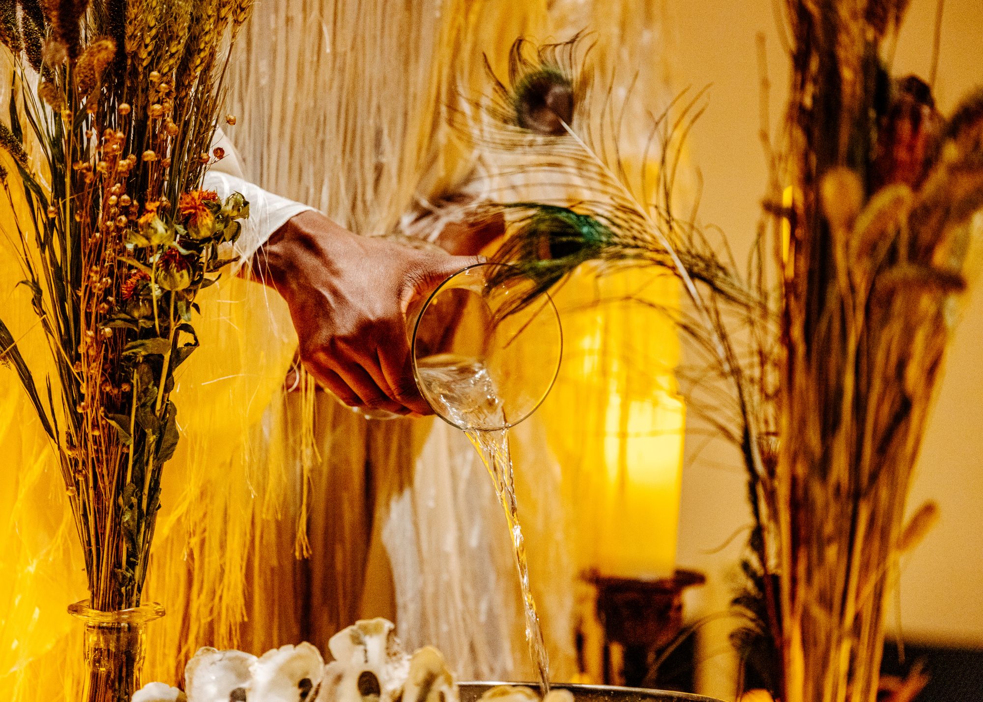 two hands pouring out water from a vase among dried plants, feathers and candles