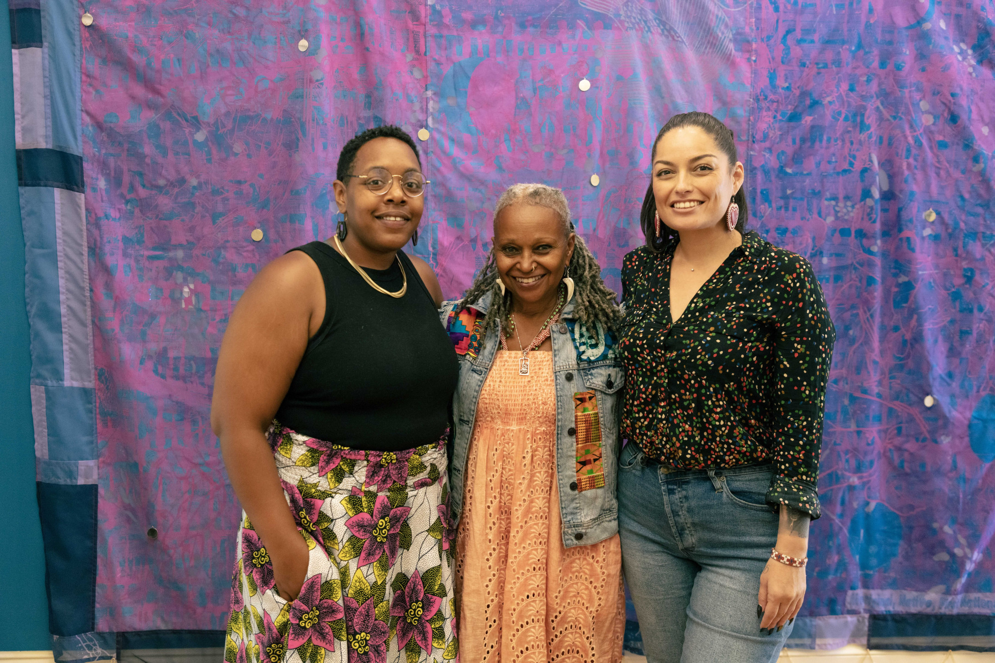 Three women standing in front of a colorful print sheet in the Simmons Center gallery.