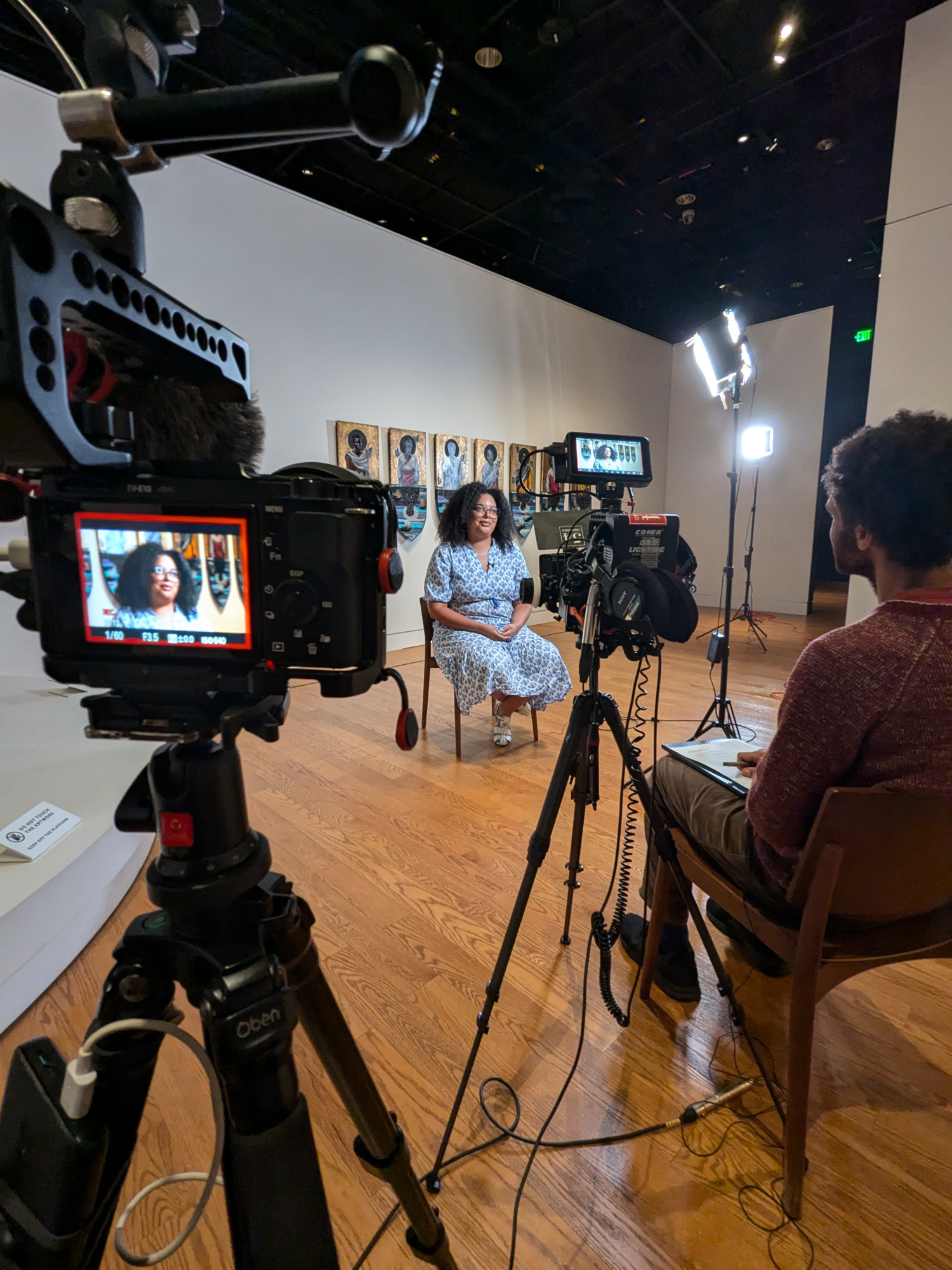 Woman seating in front of a camera set up with a man behind the cameras interviewing her.