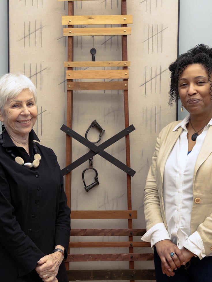 Two women stand on either side of a piece of artwork and smile at the camera