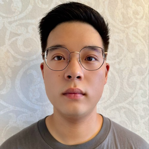 A man wearing wire-rimmed glasses looks at the camera wearing a grey shirt and standing in front of a patterned white wallpapered wall