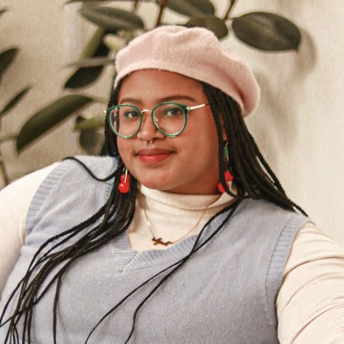 A woman wearing wire-rimmed glasses and a beret sits indoors and smiles at the camera