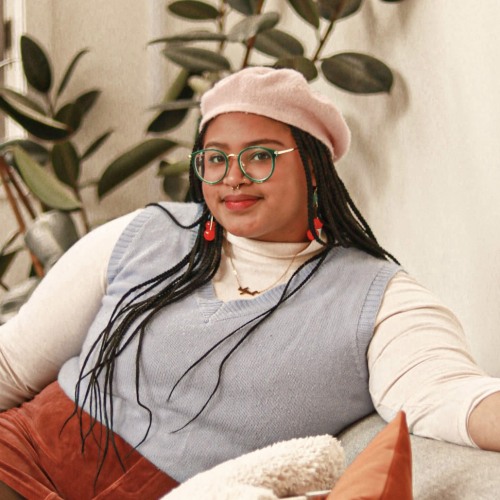 A woman with a cream beret and long braids sits on a couch and smiles at the camera