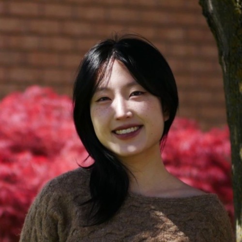 woman with straight black hair stands under a tree and smiles at the camera with a red bush in the backround
