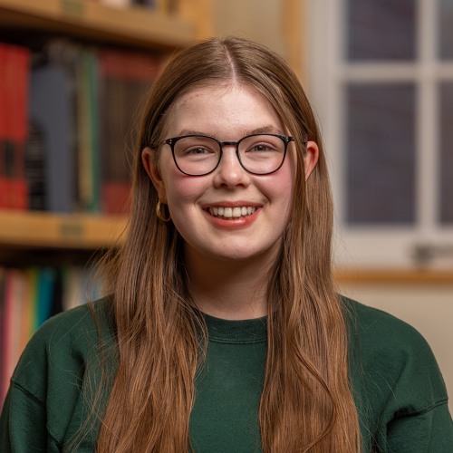 A woman with light brown hair and black rimmed glasses smiles at the camera with a book shelf in the background