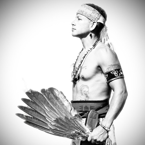 A black and white photo of a man wearing a wampum headband and holding a feather fan. Thawn Sherenté Harris