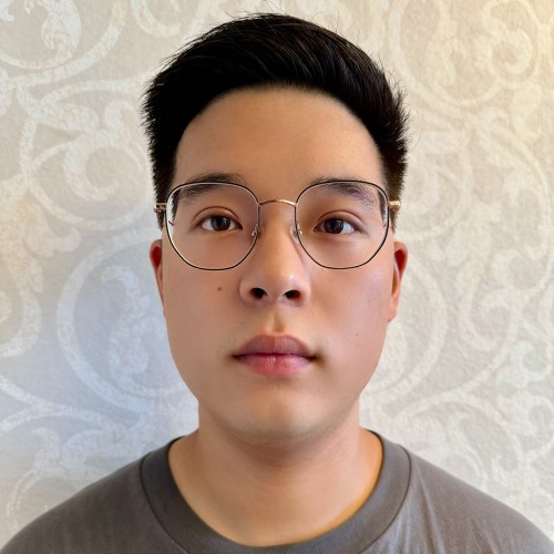 A man wearing wire-rimmed glasses looks at the camera wearing a grey shirt and standing in front of a patterned white wallpapered wall
