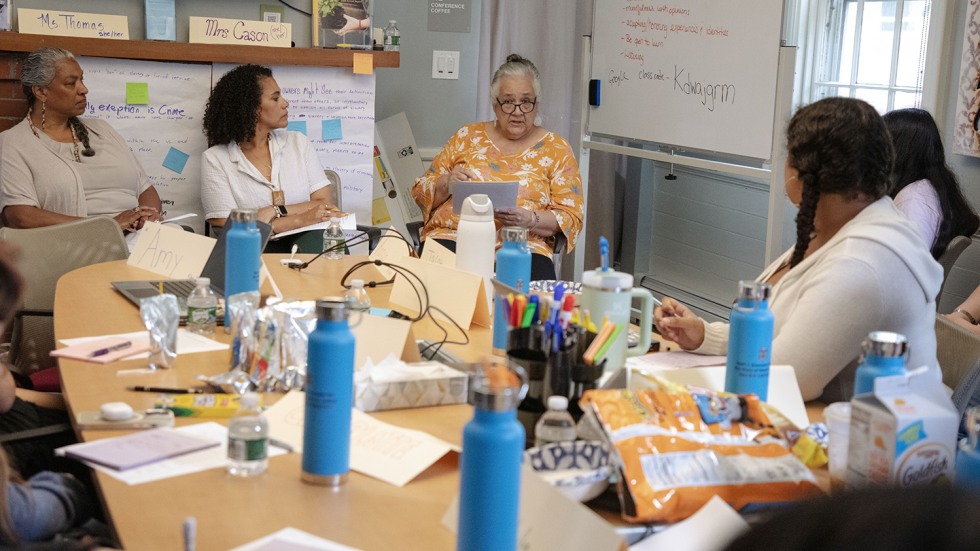 Linda Coombs sits at the front of a classroom and speaks to a classroom of high school students seated around a table
