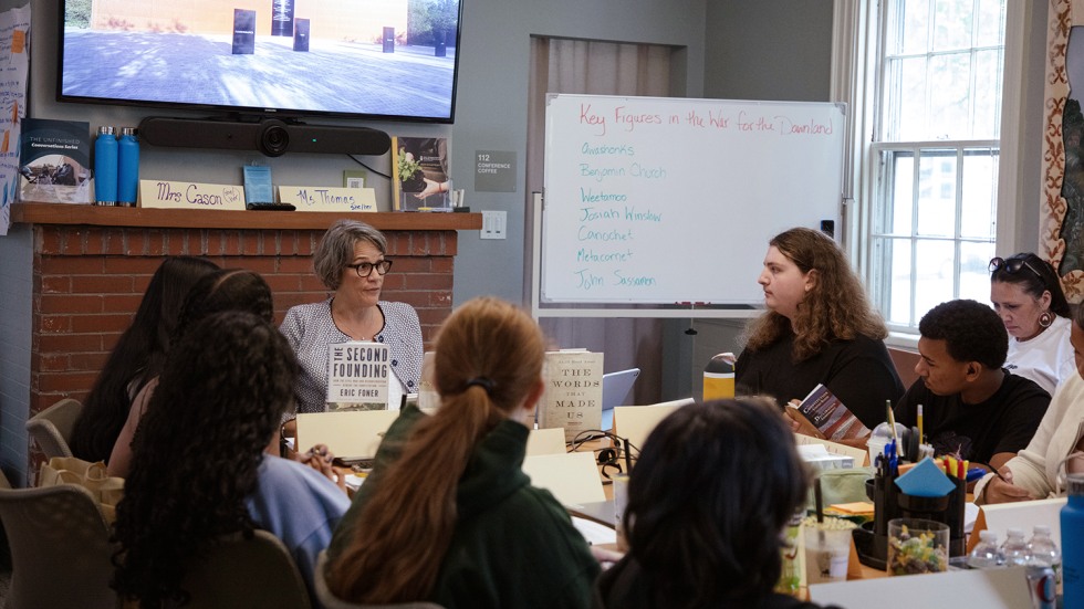 A woman site below the presentation screen and behind two books that stand upright on the table while speaking with students