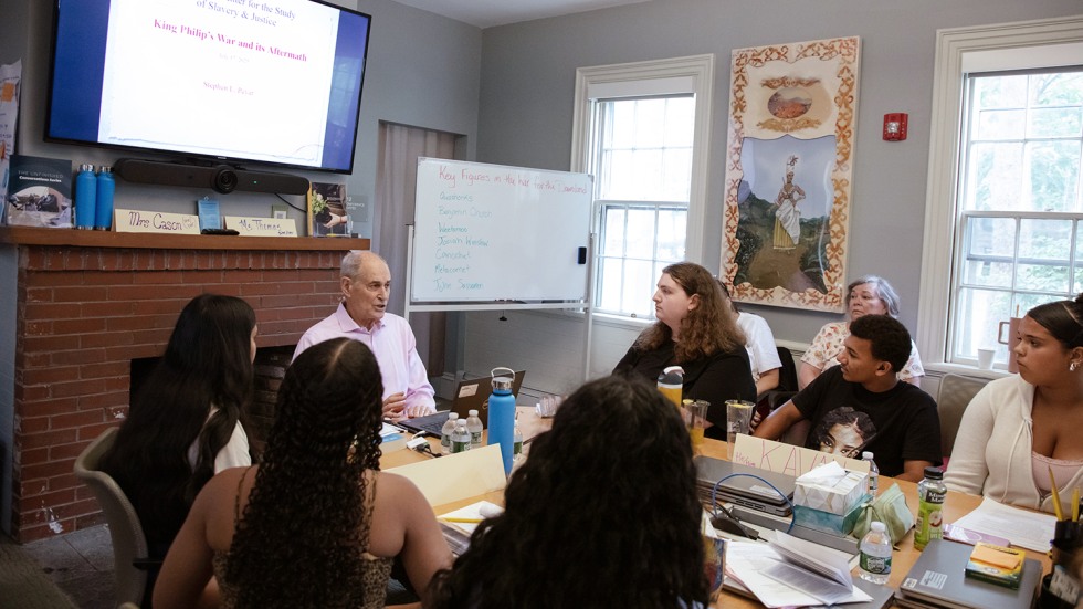 A man sits below a presentation screen and addresses students seated around a table