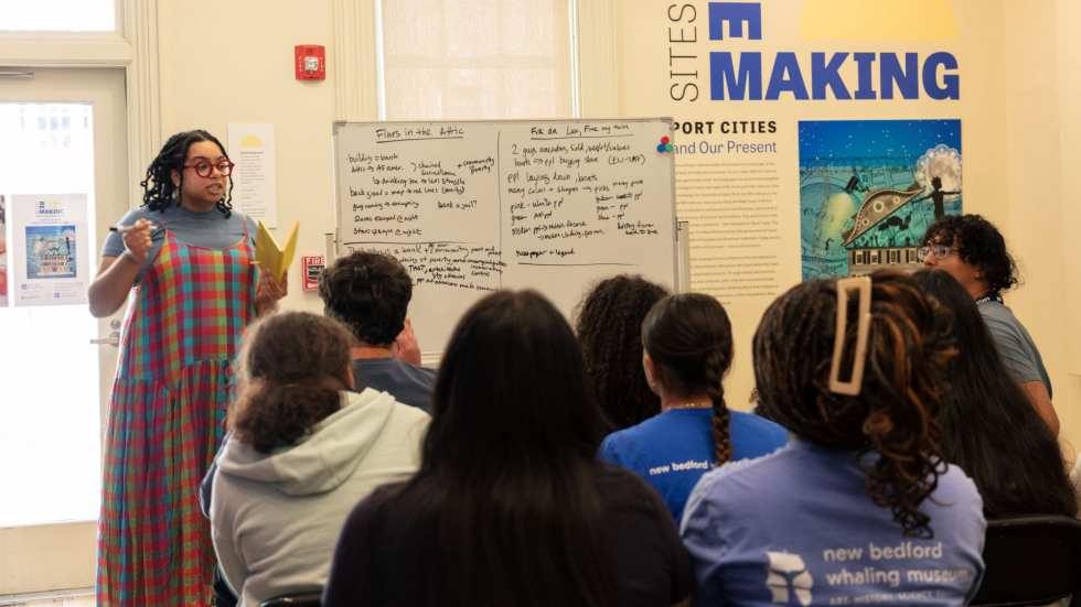 An educator stands by a whiteboard in an art gallery with several rows of students facing her.