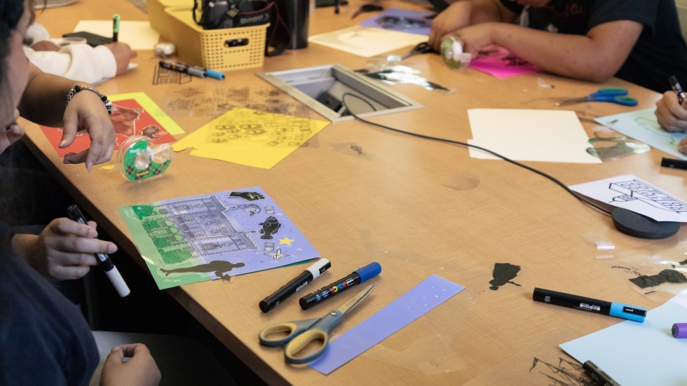 Several people work on their collages with markers and materials spread out on the table in front of them.