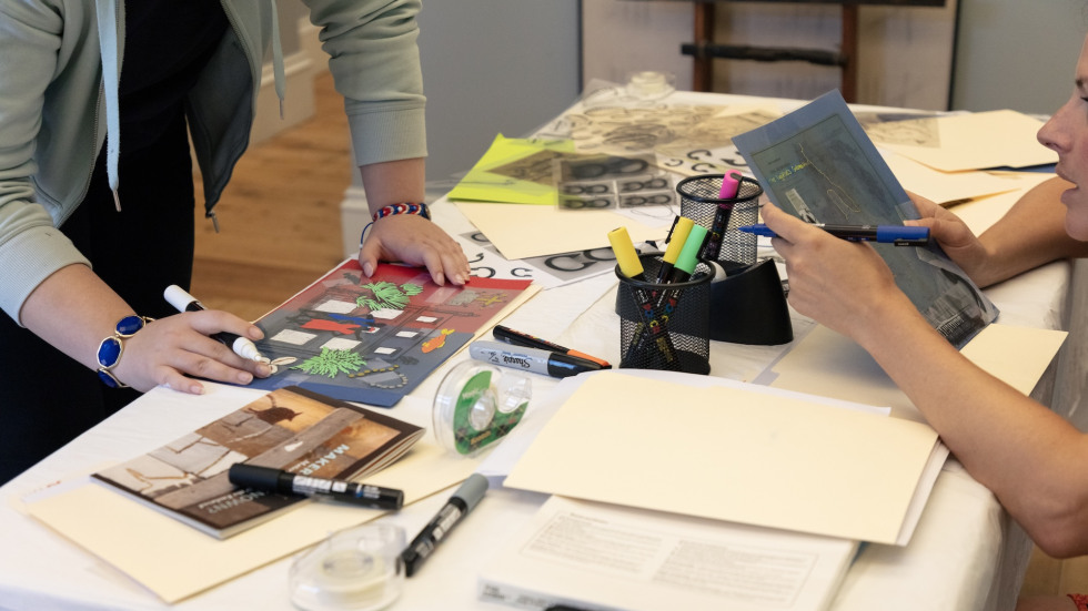 Two people work on their collages with markers and materials spread out on the table in front of them.