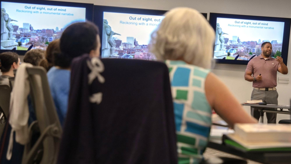 A professor stands at the front of a classroom with several people seated at tables around him. The screen in the background says Out of Sight, Out of Mind: Reckoning with a monumental narrative