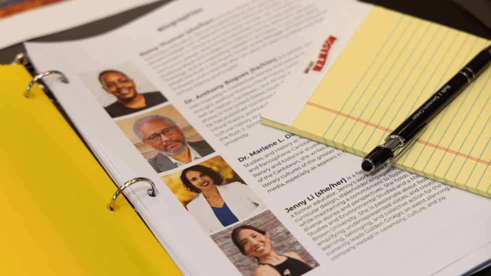 A 3-ring binder with a yellow notepad and pen sitting on top. The binder page shows 4 people’s headshots and bios.