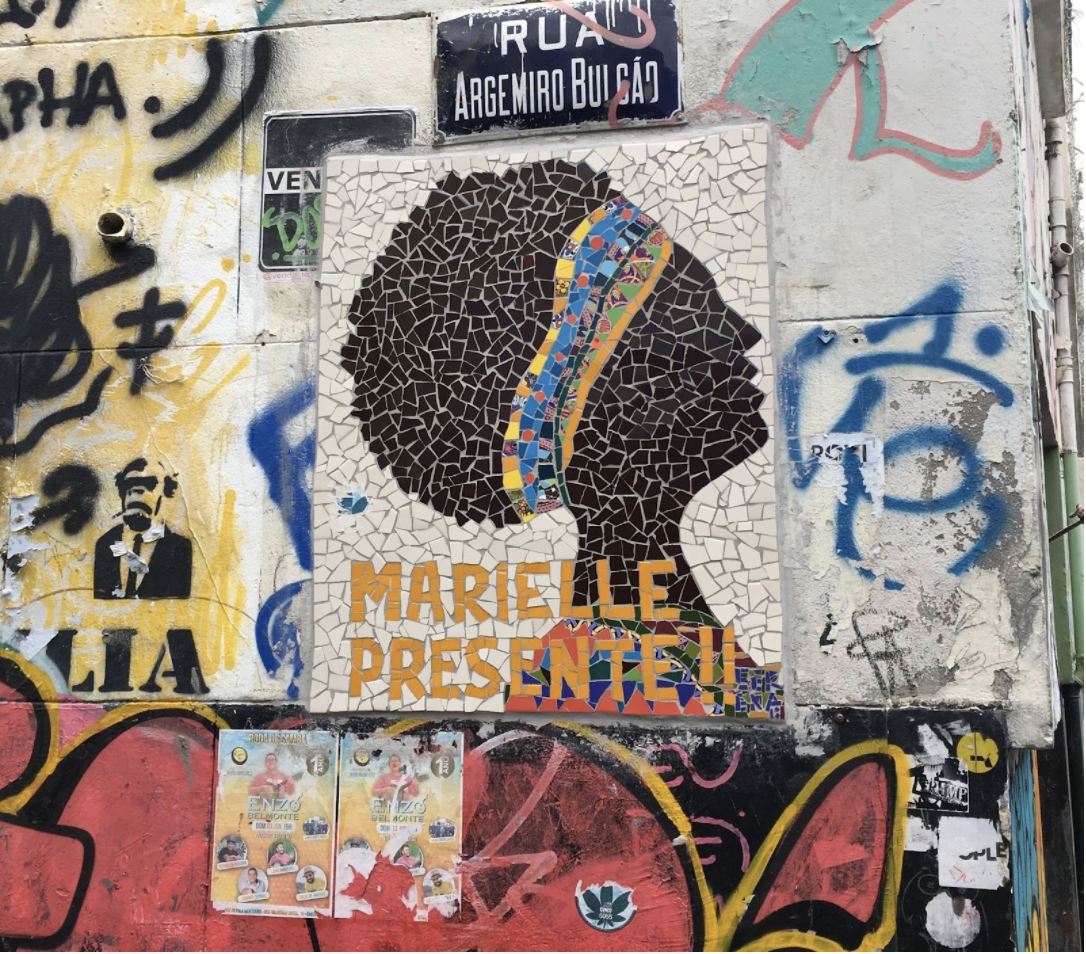 A mural of a black woman's side profile as a silhouette. She wears a colorful headband. Orange Text below reads: "Marielle Presente!!"   